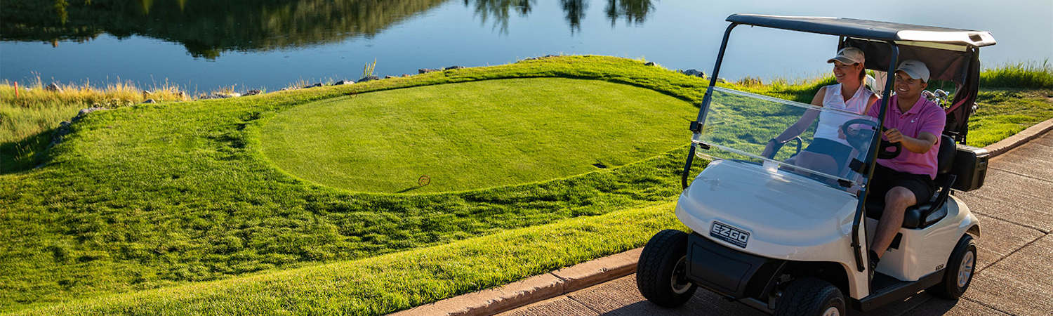 A man and woman riding in an E-Z-GO® golf cart past a putting green on a golf course.